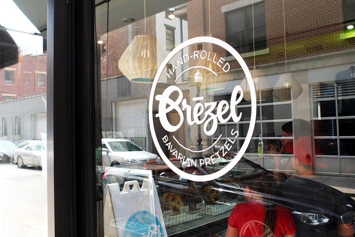 View through a glass window of a bakery shop with a round sign that reads "Hand-Rolled Brezel Bavarian Pretzels." Reflections of parked cars and street buildings are visible on the glass.