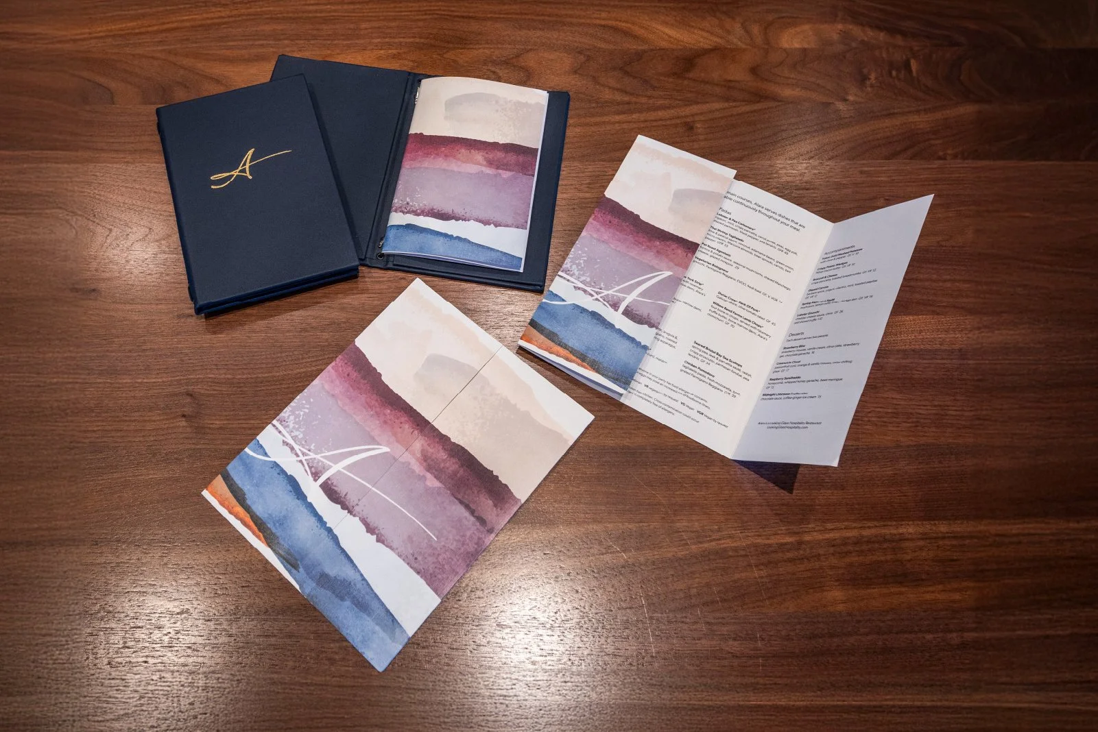 A set of brochure or menu with watercolor mountain landscape images, one is open revealing text inside, and a navy blue folder with a gold letter A on the cover is nearby, all on a wooden surface.