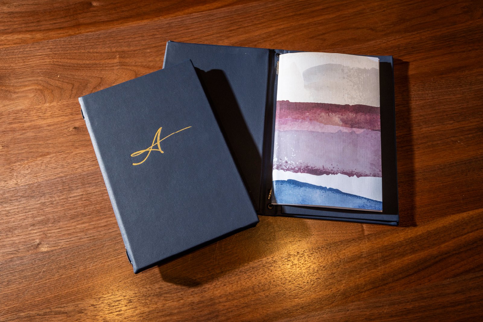 An open art portfolio with a painted landscape inside, placed on a wooden surface, with a matching closed portfolio next to it.