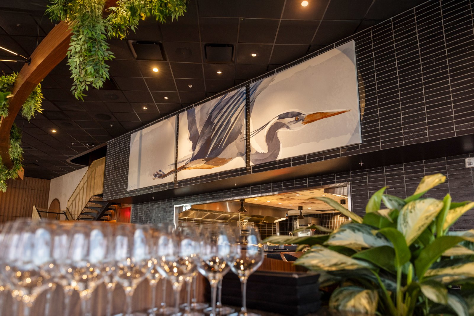 Interior of a restaurant or bar with a large mural of a heron on the wall, a row of wine glasses in the foreground, and green plant leaves partially visible in the lower right corner.