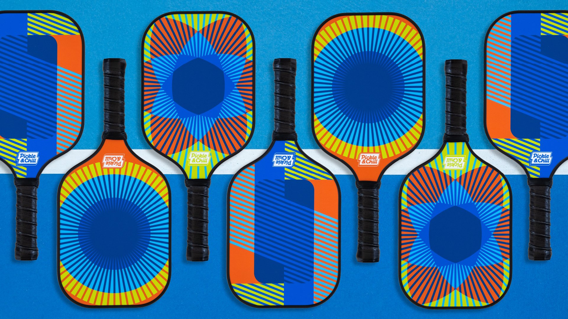 Six colorful pickleball paddles with black handles arranged on a blue surface, featuring a vibrant geometric pattern with blue, yellow, orange, and red colors.