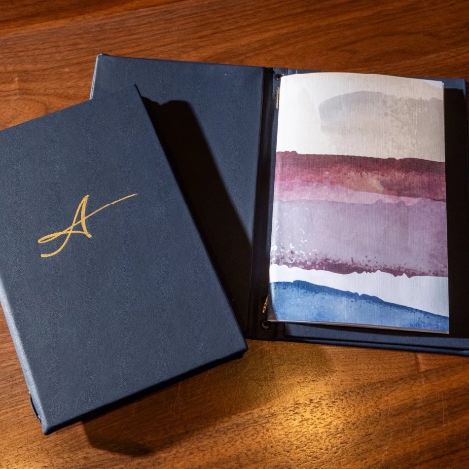 Open photo album with abstract watercolor art inside, placed on a wooden table. Next to it is a closed dark blue box with a gold letter 'A' on the cover.