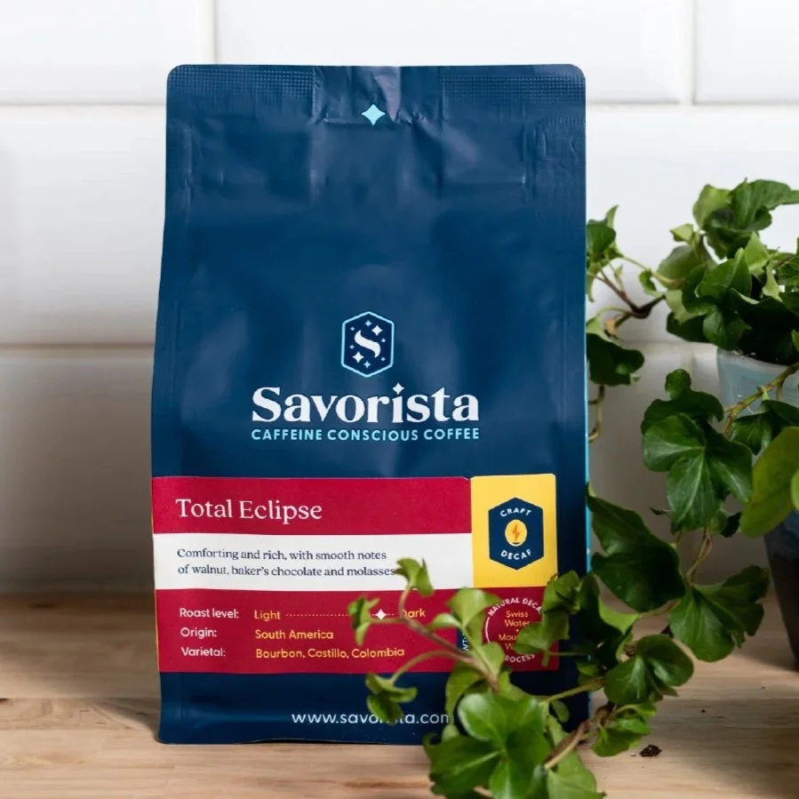 A bag of Savorista Total Eclipse coffee, labeled as caffeine conscious, with notes of walnut, chocolate, and molasses, placed next to a green potted plant.
