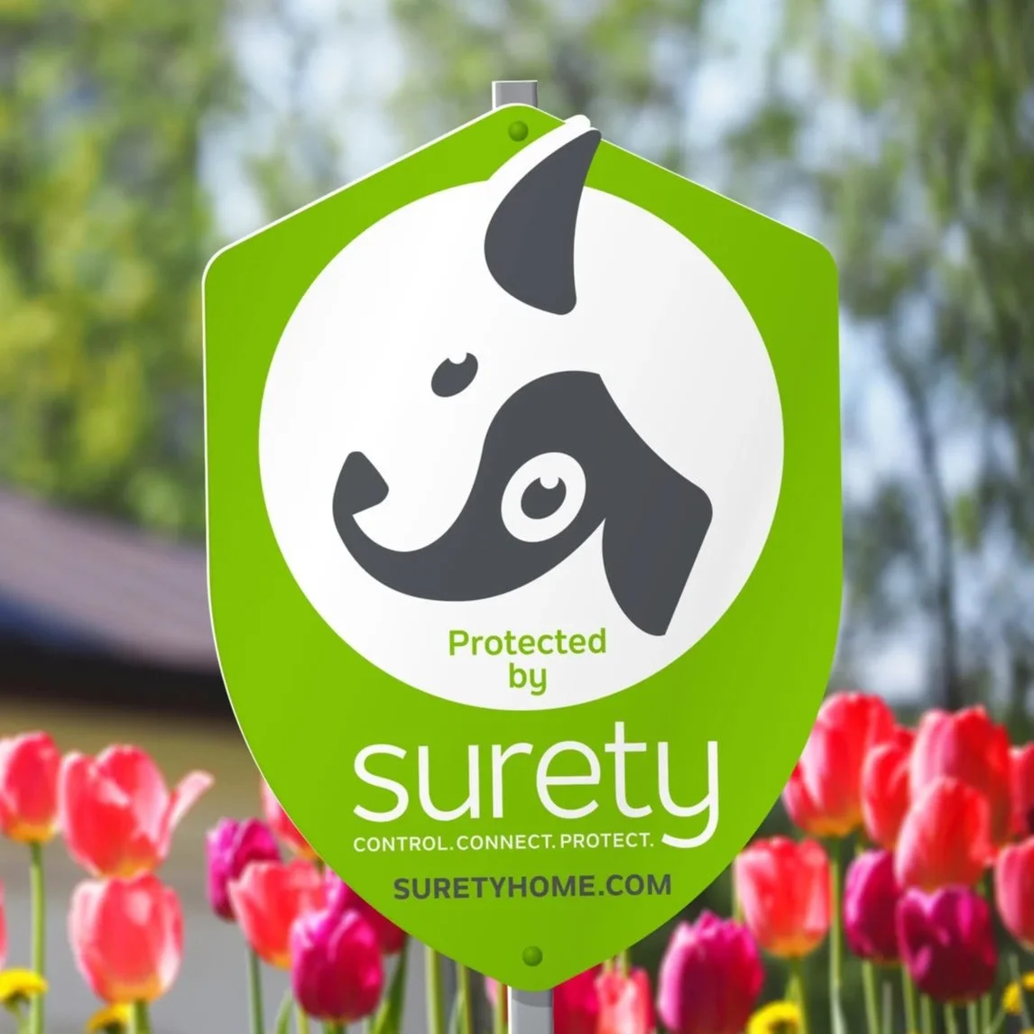 A green pet protection sign with a black and white stylized dog logo, stating "Protected by surety" and the website suretyhome.com, with pink and yellow tulips in the background.