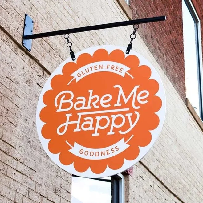 Round sign with orange border and white background, hanging outside a brick building, displaying the words 'Bake Me Happy' with phrases 'Gluten-Free' at the top and 'Goodness' at the bottom.
