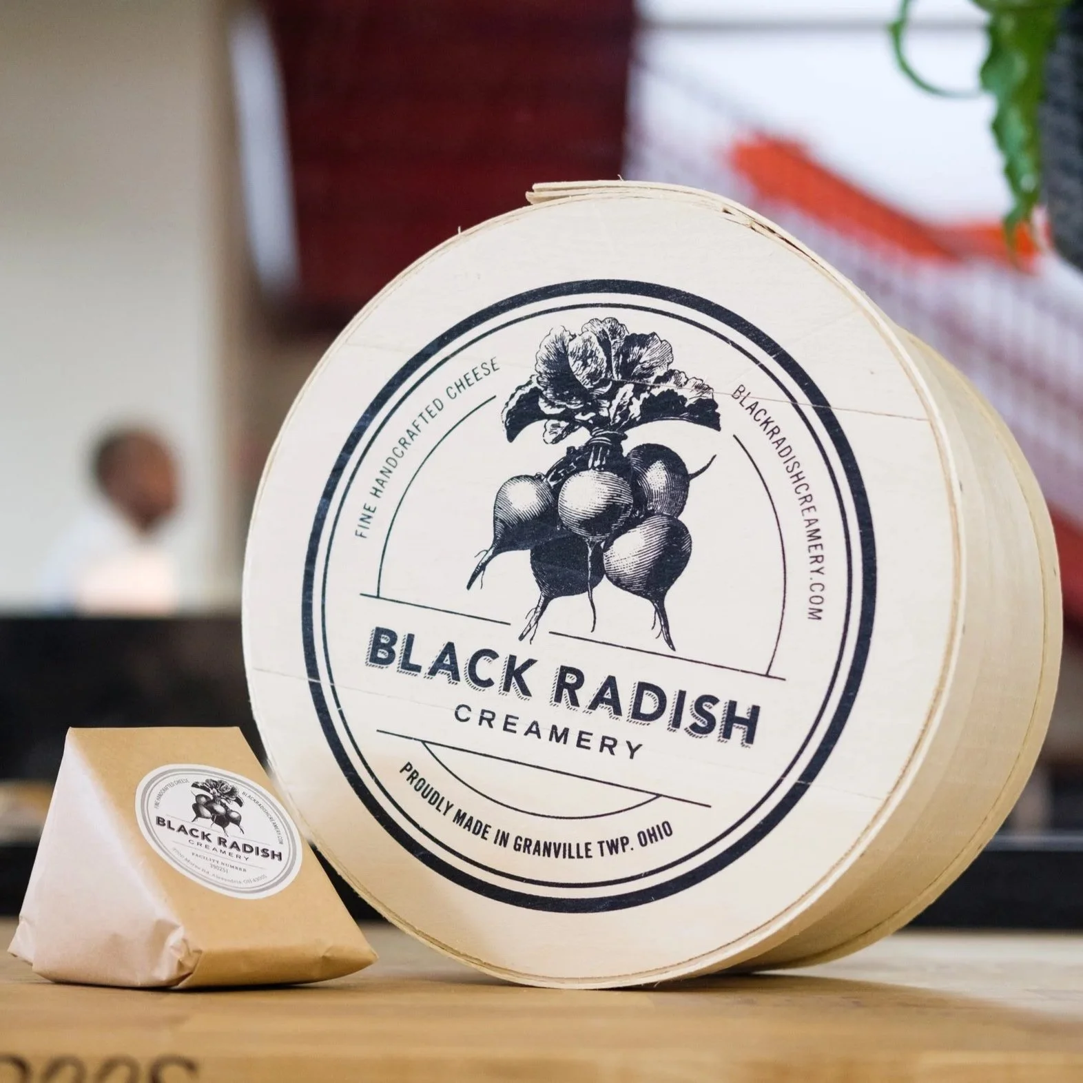 A round container of Black Radish Creamery cheese with a label featuring a black and white illustration of a radish and the brand name. A paper bag with the same logo is in front of it, and a person blurred in the background.