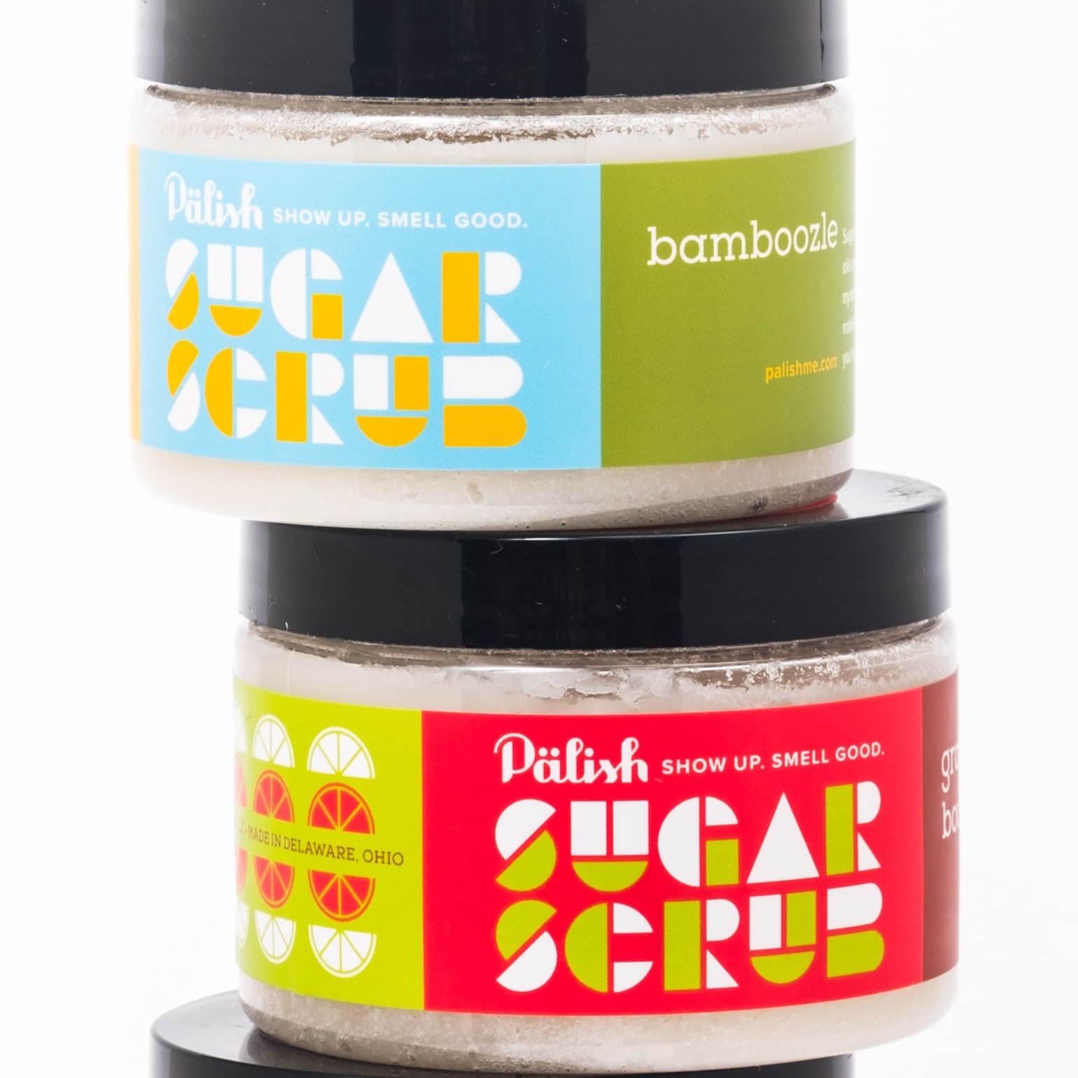 Close-up of two containers of flavored sugar scrub, one with a blue label and one with a red label, stacked on top of each other on a black stand. The labels include colorful citrus fruit graphics and text.