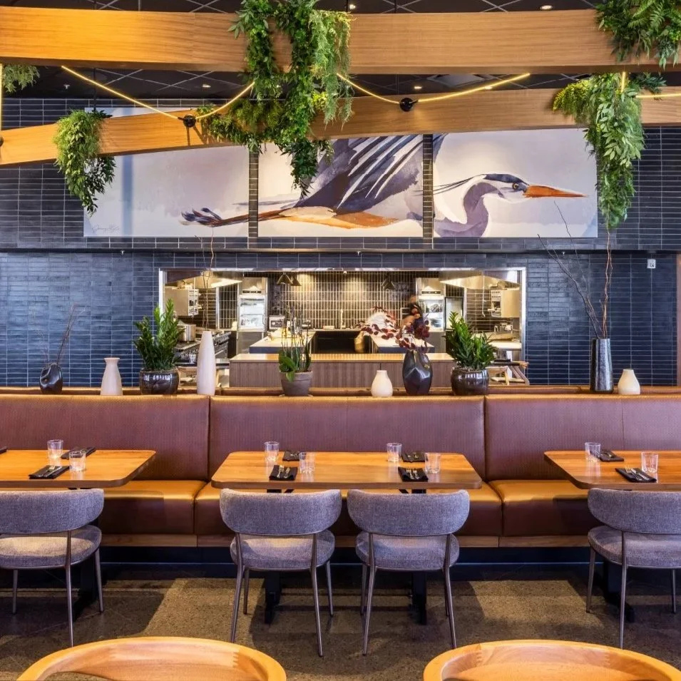 Interior of a modern restaurant with a mural of a heron on the wall, decorated with hanging plants, vases, and potted plants on a shelf, with tables set for dining.