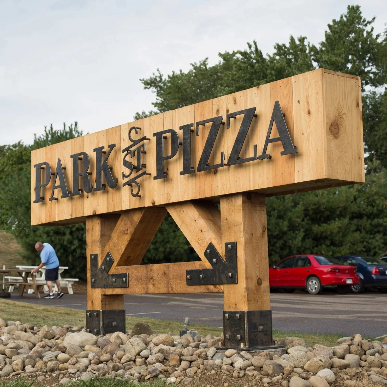 A wooden sign reading 'Park & DŻA PIZZA' with decorative metal elements, situated on rocks near a parking lot with a red car and a man at a picnic table in the background.