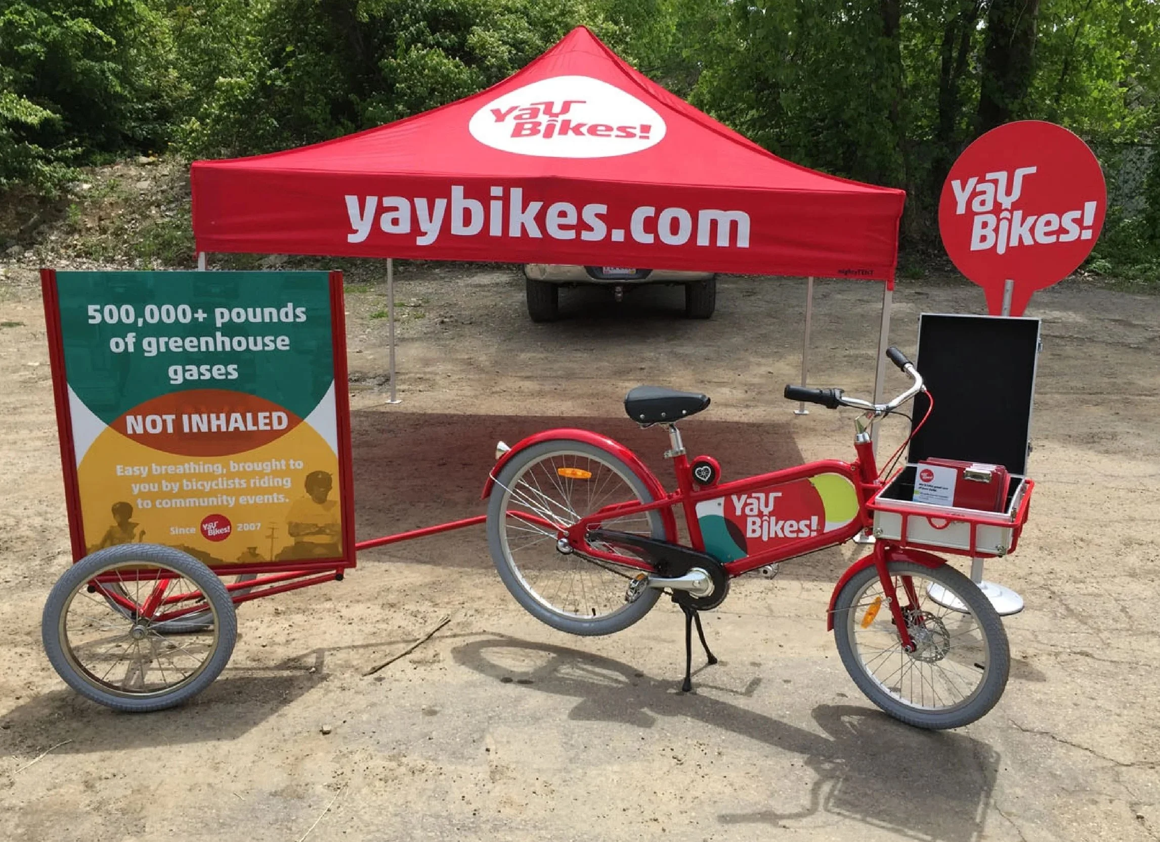 A red bicycle with a cargo box on the front, branded with "Yay Bikes!", parked in front of a red canopy tent with the same branding. There is a signboard to the left promoting the environmental benefits of biking, and a circular sign to the right als