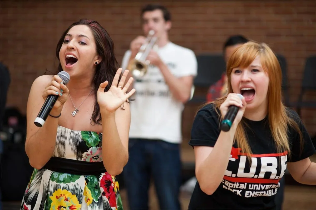 Two women singing into microphones and a man playing trumpet in the background during a musical performance.