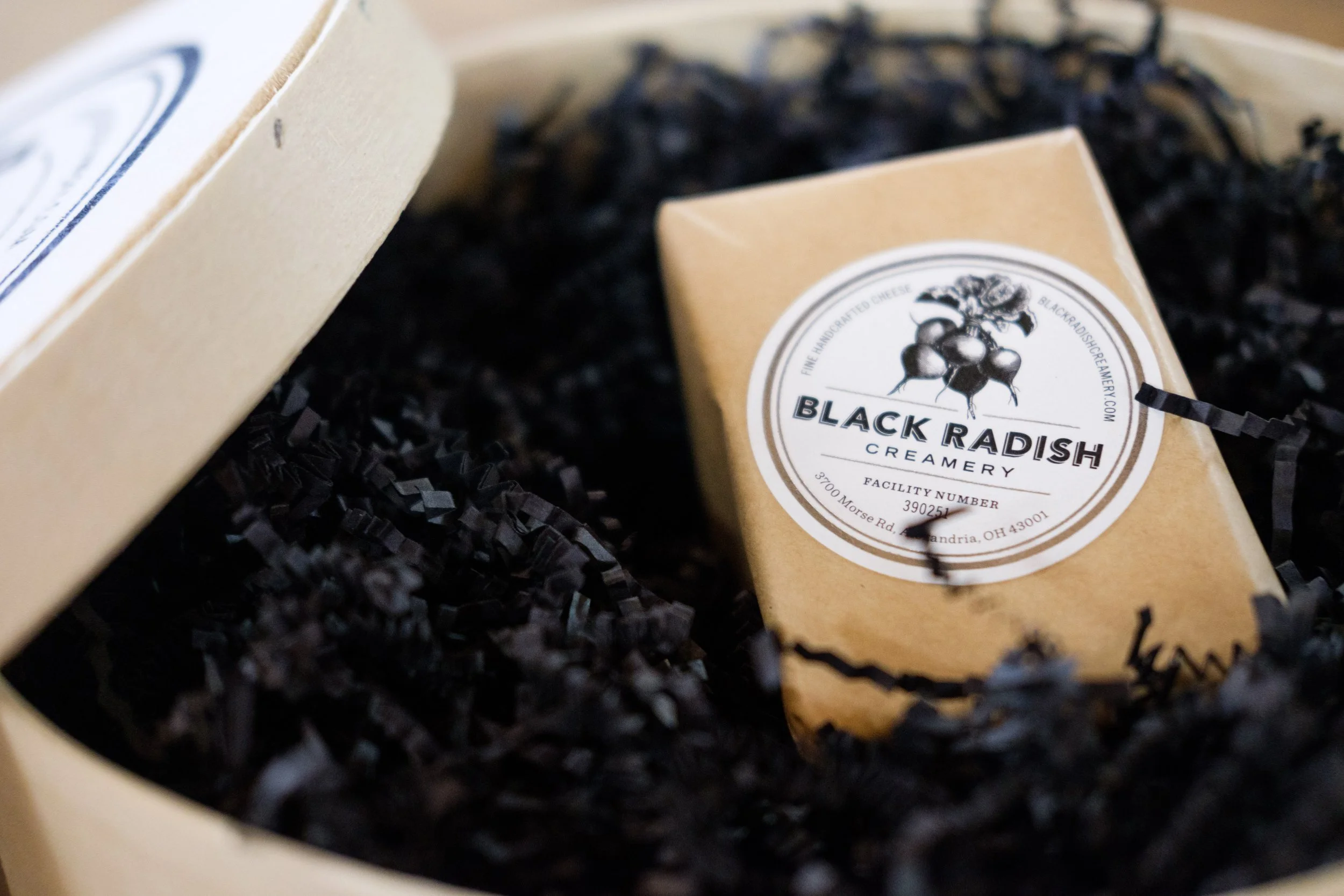 Close-up of a package of Black Radish Creamery cheese in a box filled with black shredded paper.