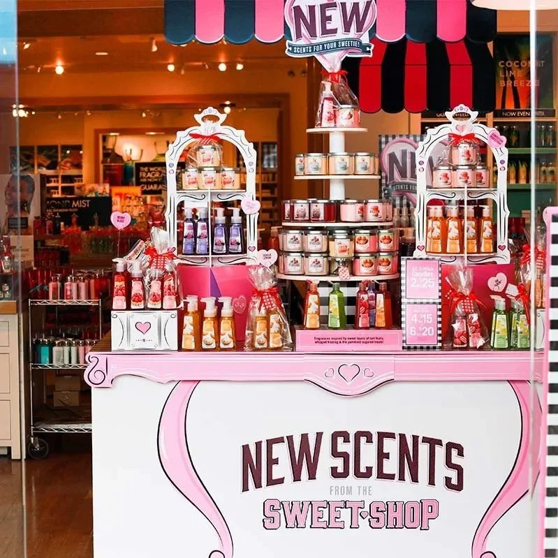 Display of scented products from the New Scents shop, featuring bottles and jars with decorative pink and red ribbons and hearts, arranged on a pink and white stand with a sign promoting new scents.
