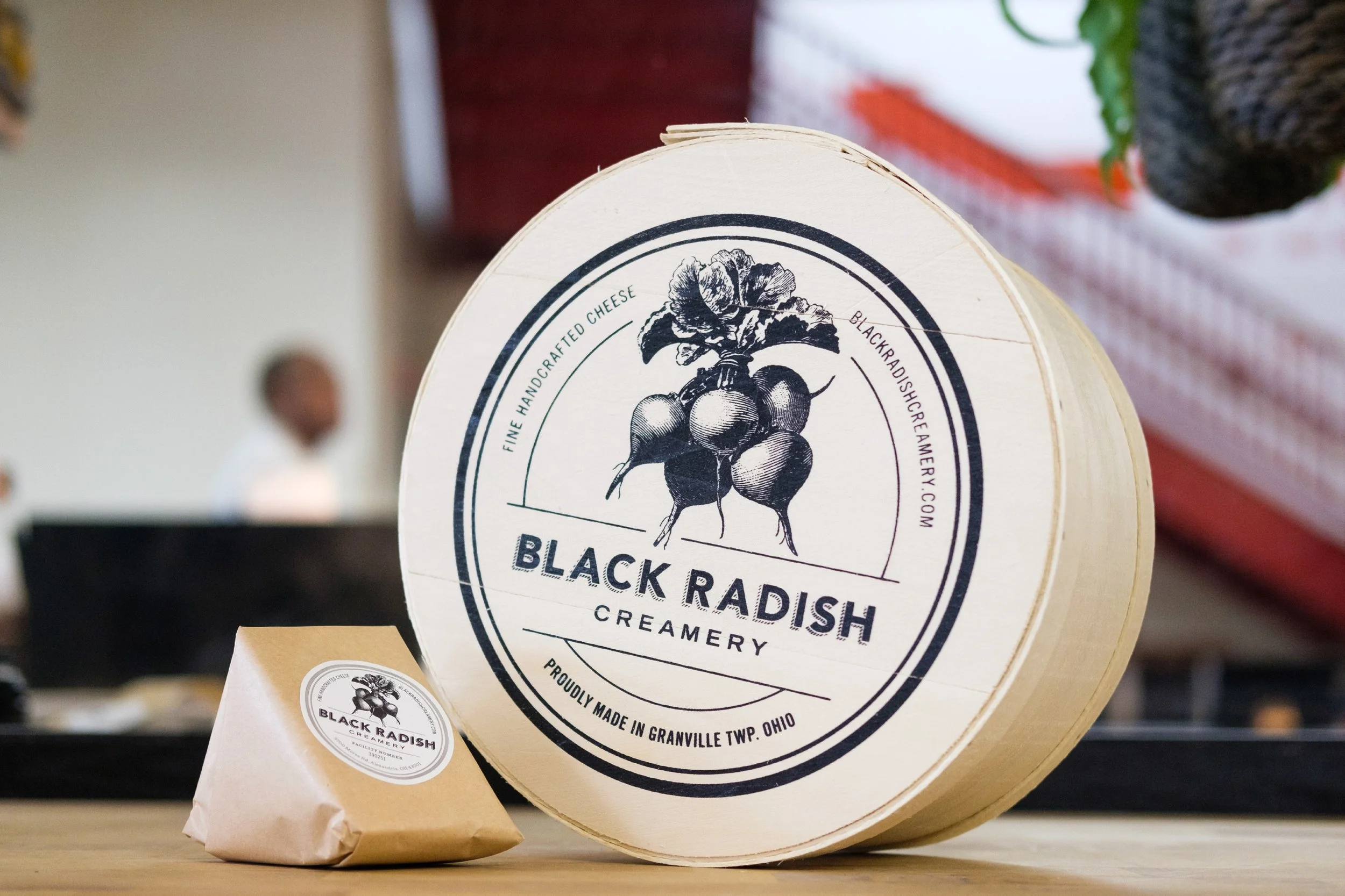 A round wooden container with a label featuring a black radish illustration, containing black radish creamery products, situated on a wooden surface.