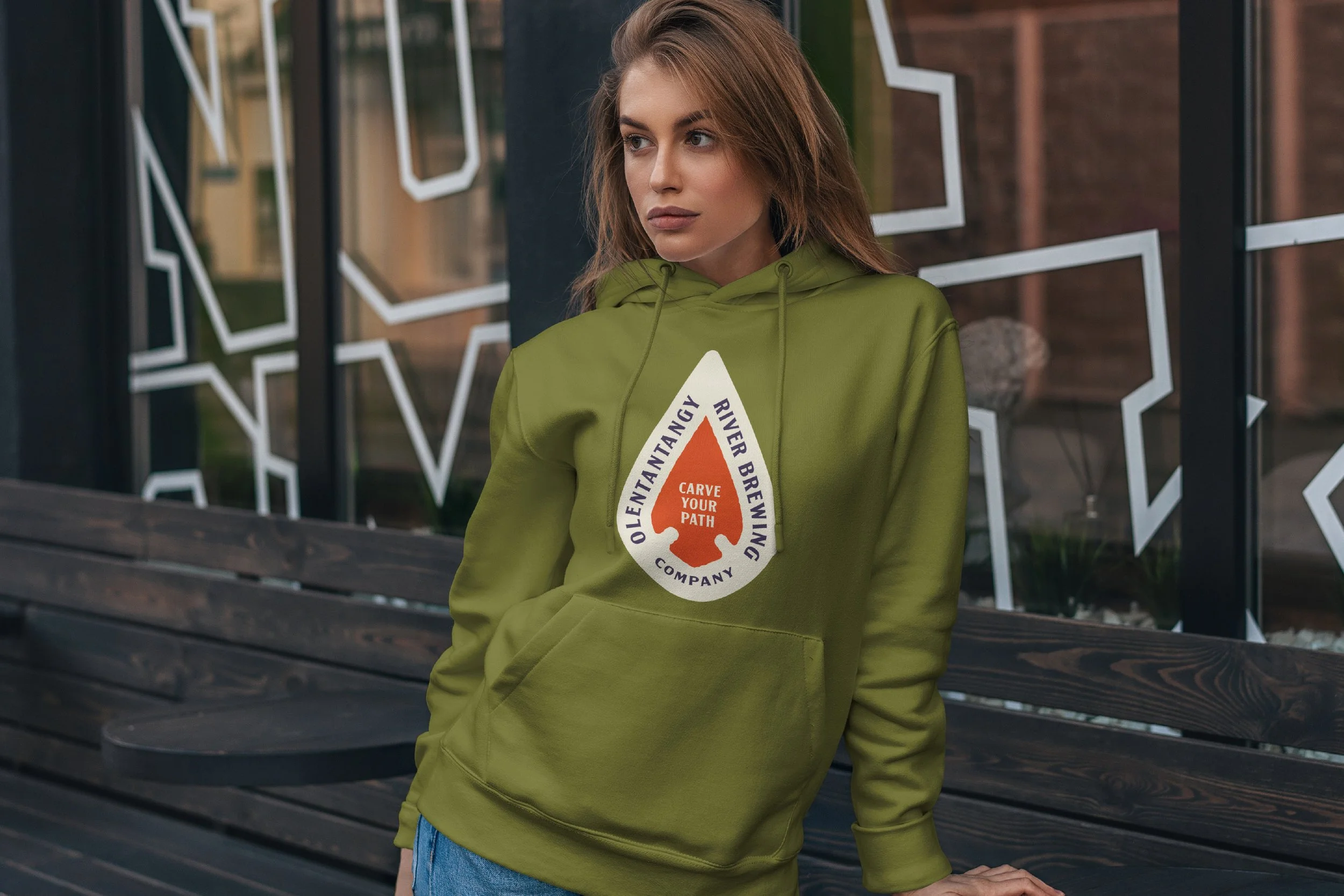A young woman with brown hair wearing a green hoodie with a logo that says 'Olen Tbeantangy River Brewing Company' and 'Carve Your Path' inside an orange triangle. She is standing outdoors near a black wooden railing and a glass wall with white geome