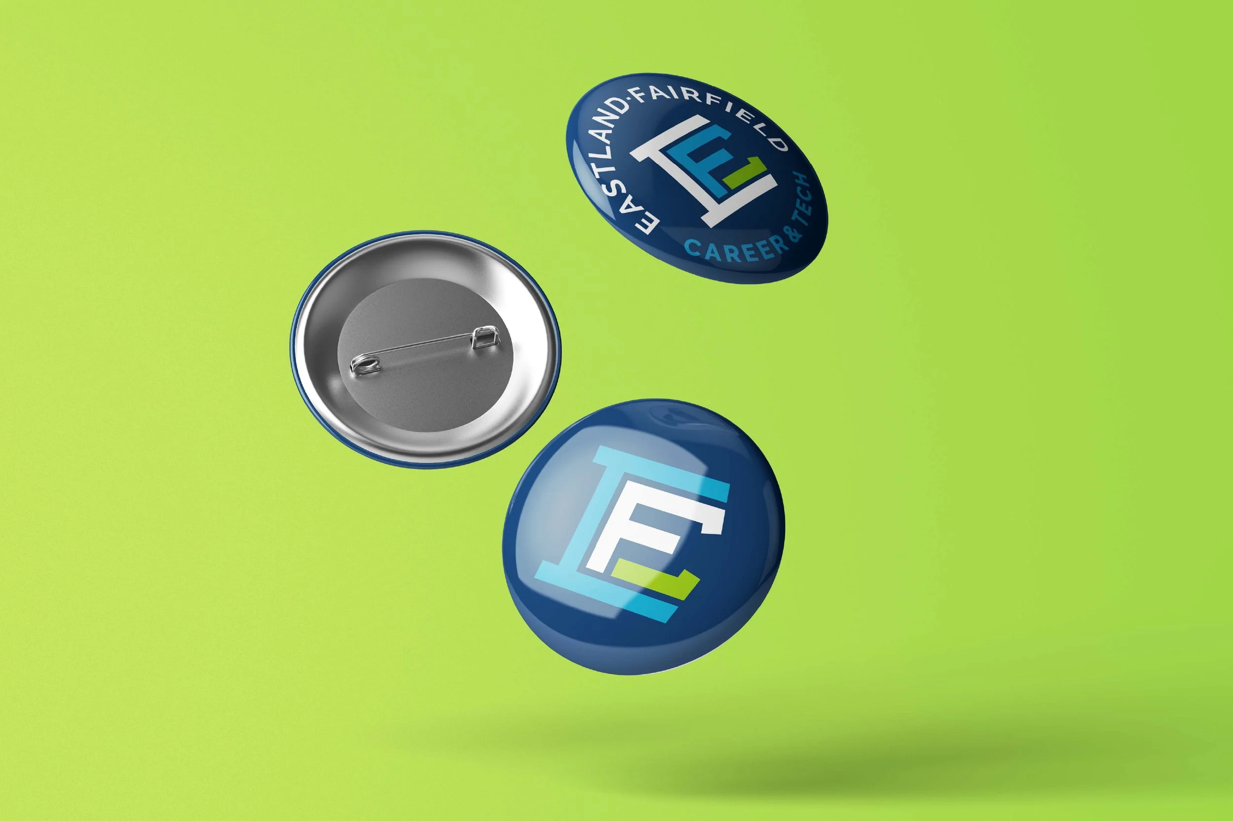 Back of a round pin button showing safety pin attachment, with three additional round buttons showing blue and green logos and text about Eastland-Fairfield Career & Tech.