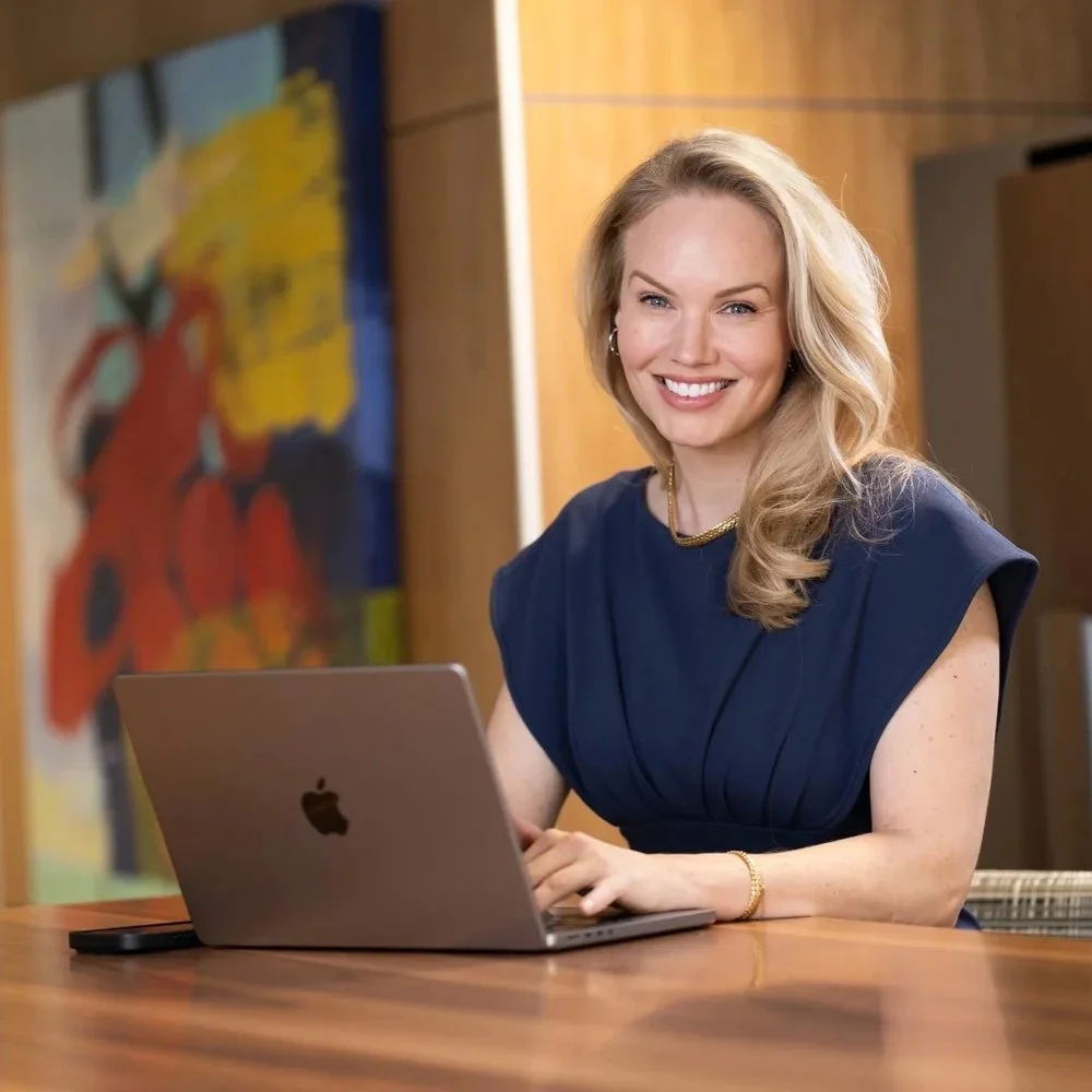 A smiling woman with blonde hair working on a laptop at a desk in a modern office.