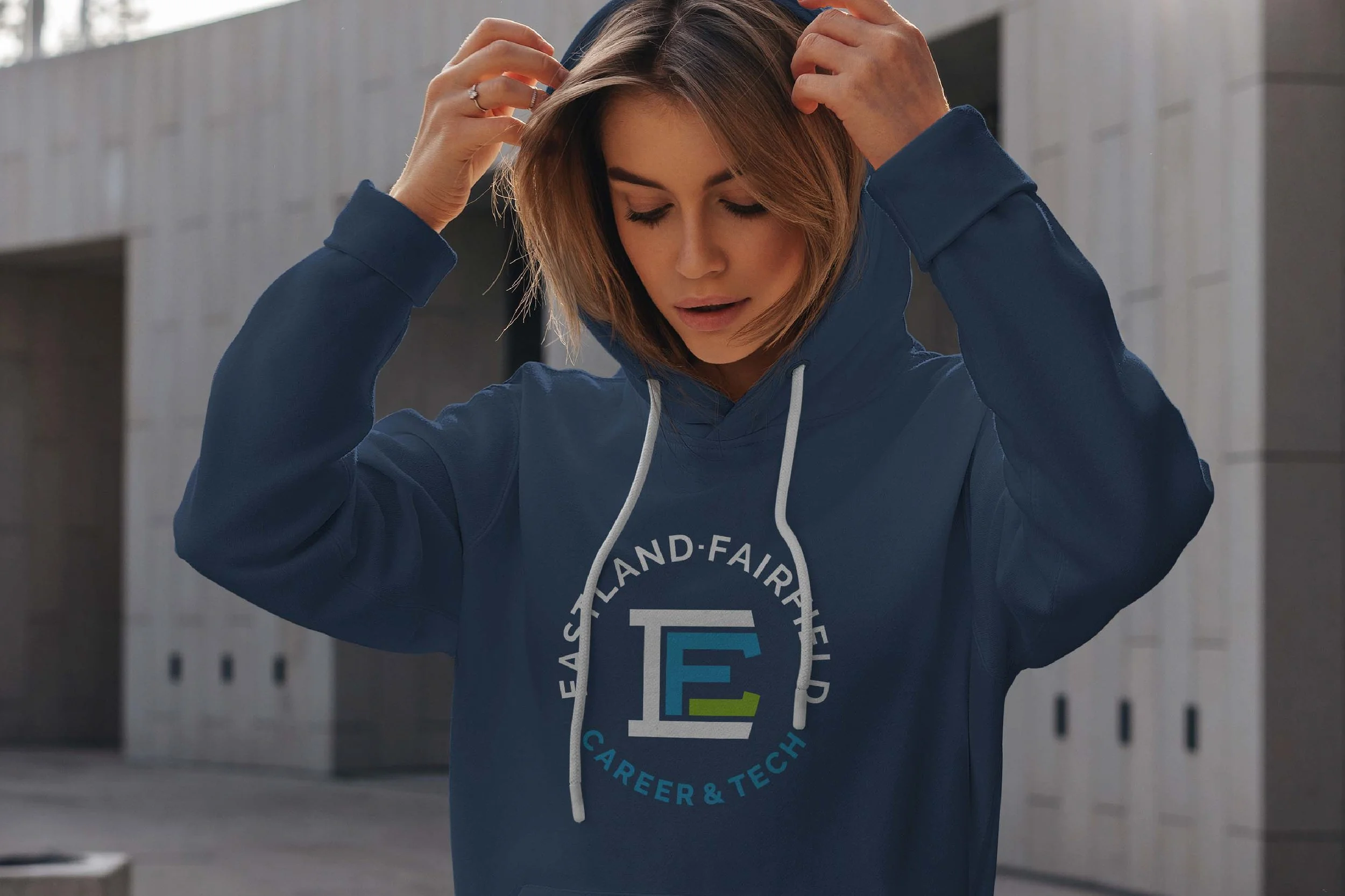 Young woman in a navy blue hoodie adjusting her hair, standing outdoors near a building with concrete walls.