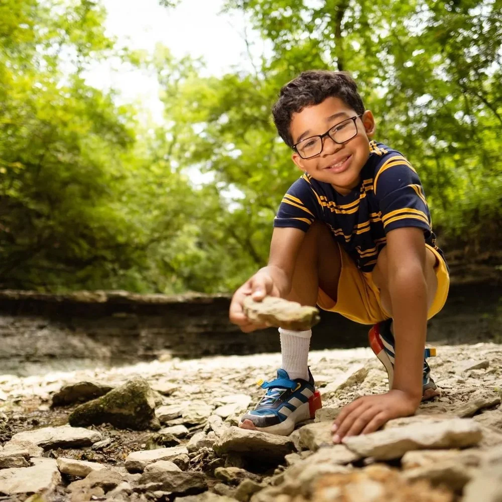 A young boy with glasses, a striped navy and yellow shirt, yellow shorts, and sneakers, crouching on rocky ground in a forest, holding a rock and smiling at the camera.