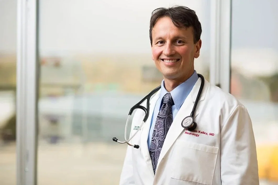 A smiling male doctor in a white coat with a stethoscope around his neck, standing in front of a window.