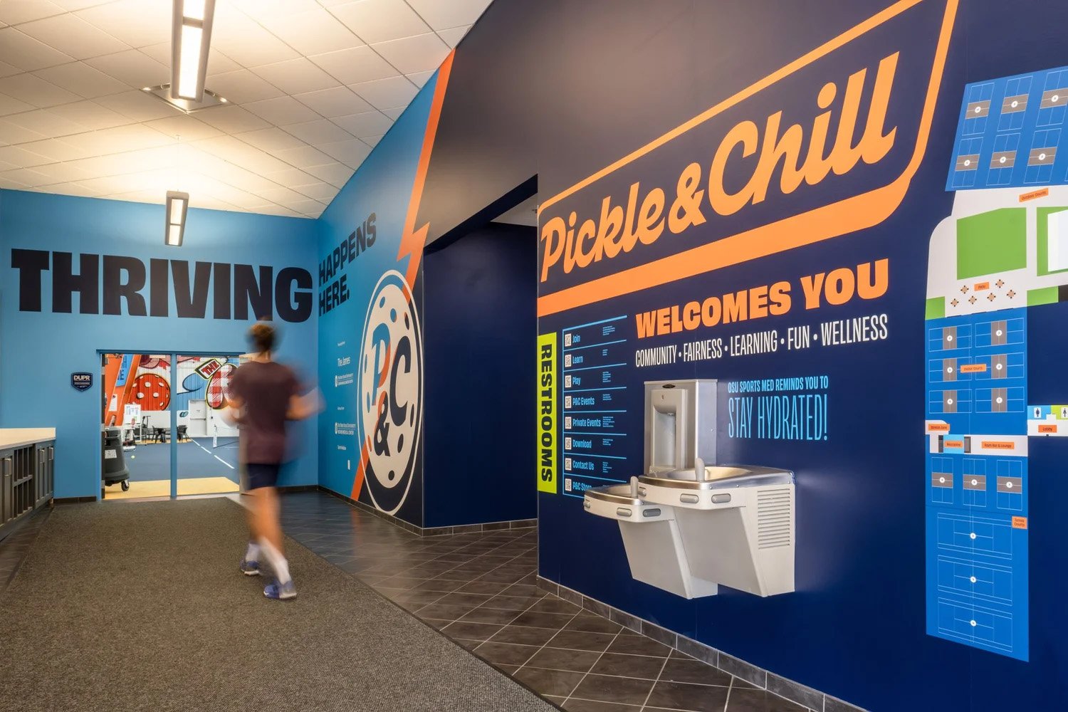 Indoor sports facility hallway with blue walls, sports-themed murals, and signage for pickle and chill activities, featuring a water fountain and a person jogging.