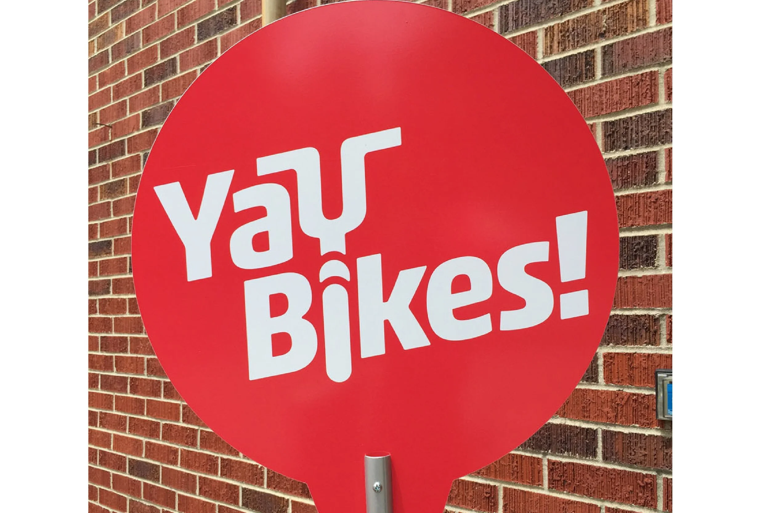 A red circular sign on a pole with white text saying 'No Bikes' in front of a brick wall.