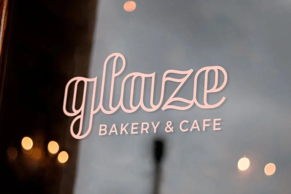 Sign for Glaze Bakery & Cafe with pink lettering on a glass window, reflecting lights and a cloudy sky outside.