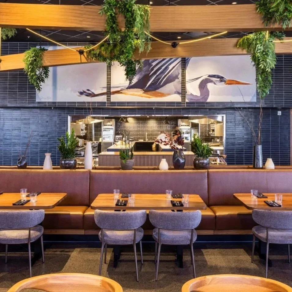 Interior of a restaurant with a mural of a heron on the wall above a kitchen pass-through window. There are wooden tables with glasses and black napkins, gray chairs, and a brown upholstered booth. Decorative vases with plants and greenery hanging from the ceiling are also visible.