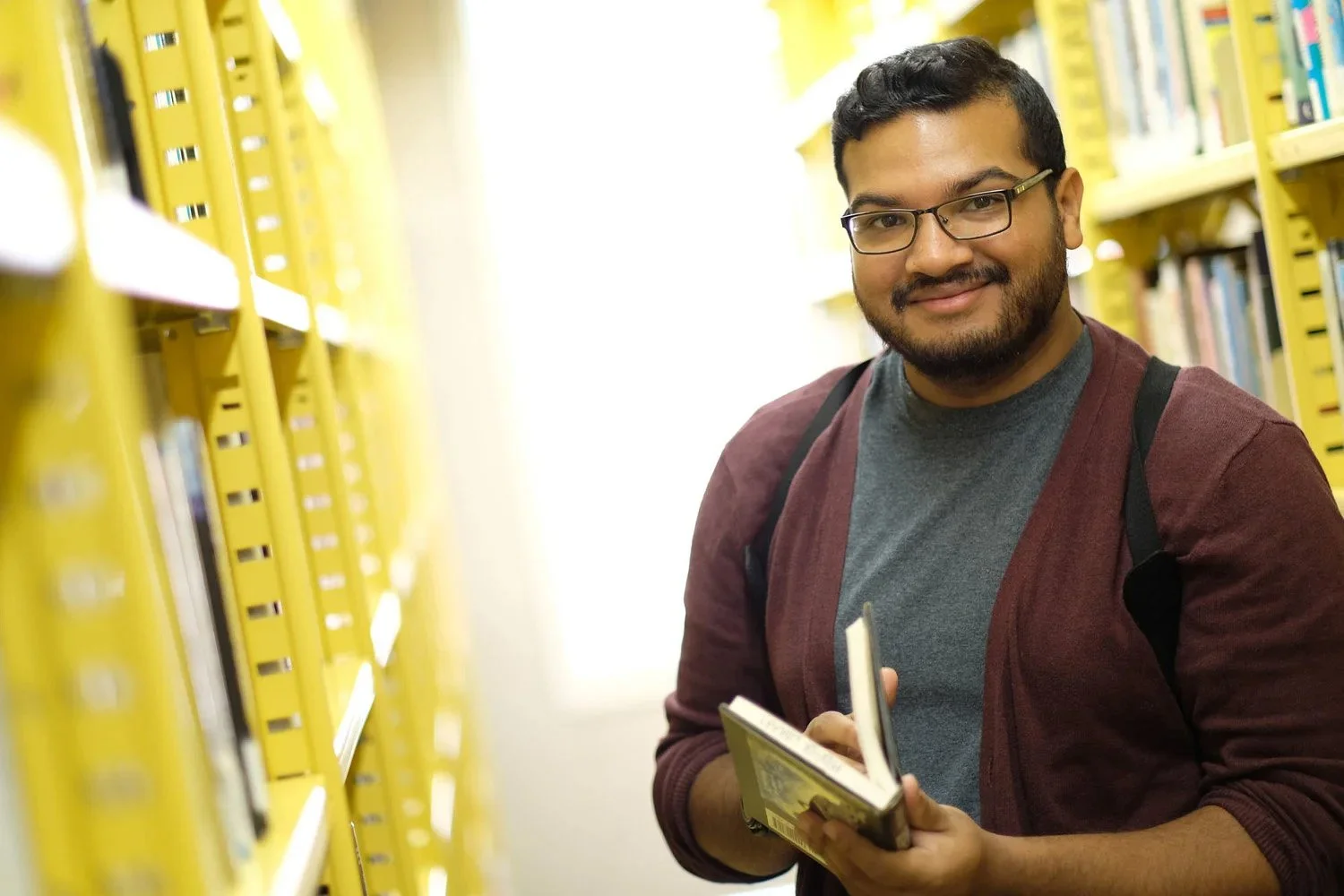 A young man with glasses and a beard, smiling while holding a book, standing in a library aisle with yellow shelves filled with books.