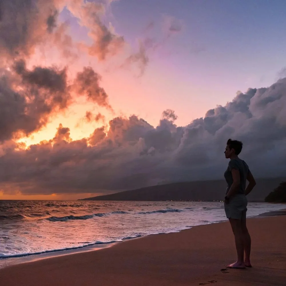 A woman standing on a beach during sunset, with clouds in the sky and ocean waves in the foreground.