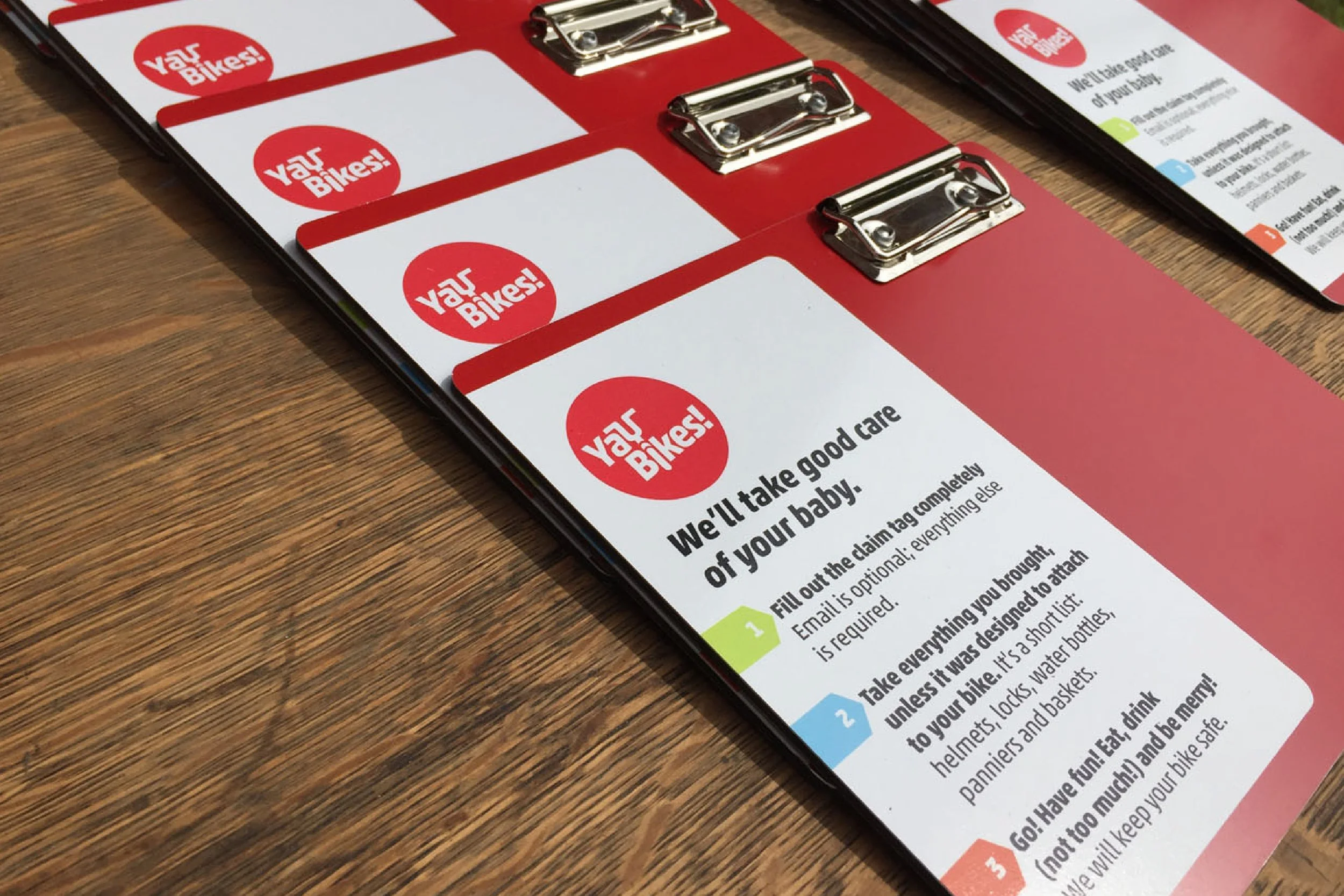 A close-up view of a red and white clipboard on a wooden table, displaying multiple identical flyers with the logo and name "Your Bikes!" and a part of an informational pamphlet next to it.