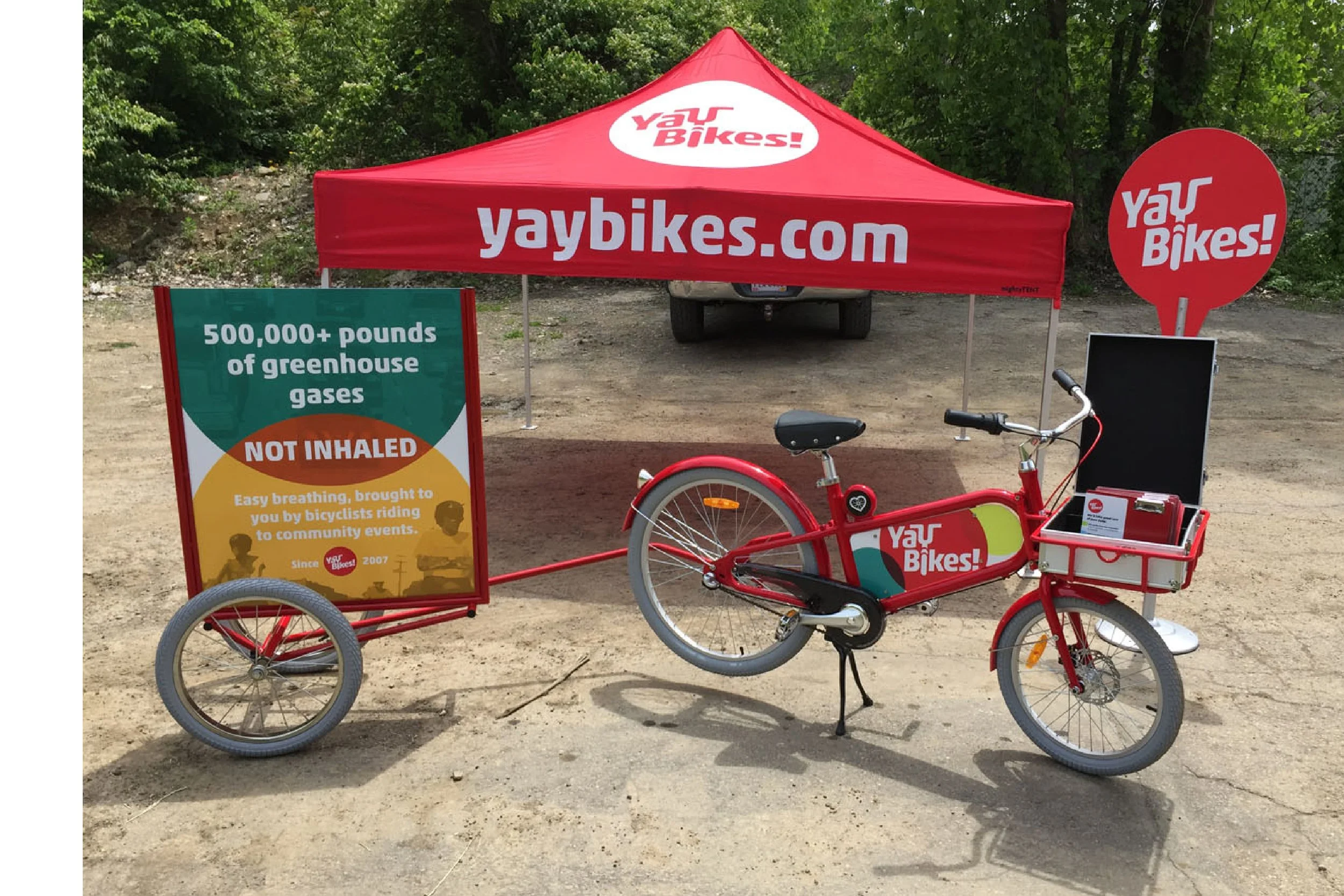 A red bicycle with a cargo box on the front, branded with "Yay Bikes!", parked in front of a red canopy tent with the same branding. There is a signboard to the left promoting the environmental benefits of biking, and a circular sign to the right als