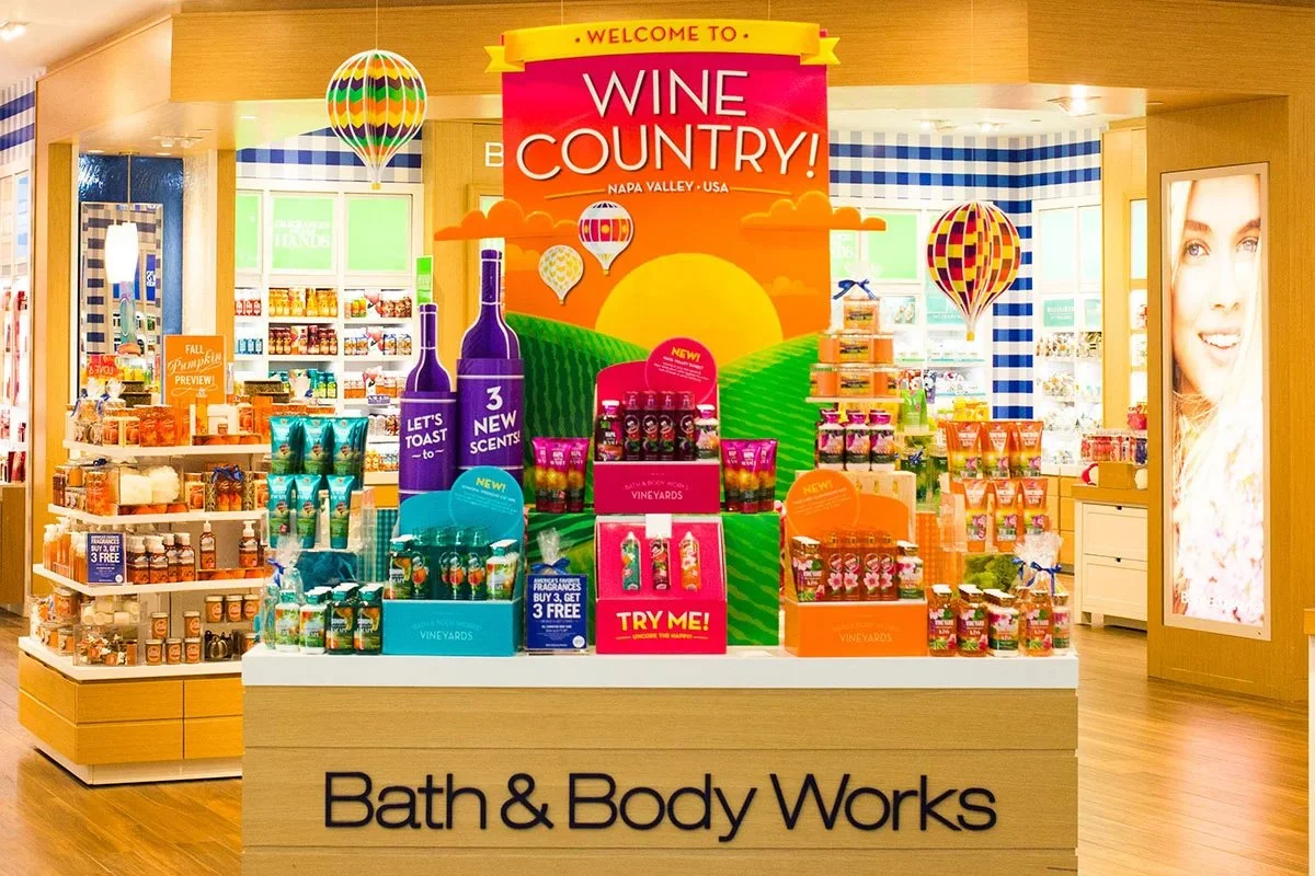 In-store display at Bath & Body Works with a banner that reads 'Welcome to Wine Country! Napa Valley, USA.' The display promotes wine-scented products with colorful bottles, signs, and decorations resembling wine bottles and hot air balloons.
