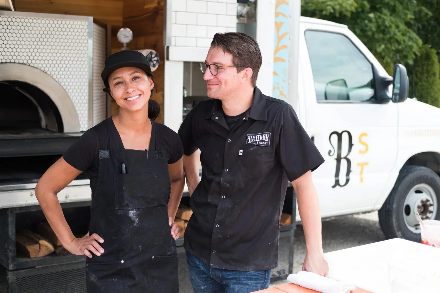 Two people, a woman and a man, standing in front of a food truck, smiling at each other. The woman wears a black hat and apron, and the man wears a black shirt with a name tag that reads "Bakker Street."