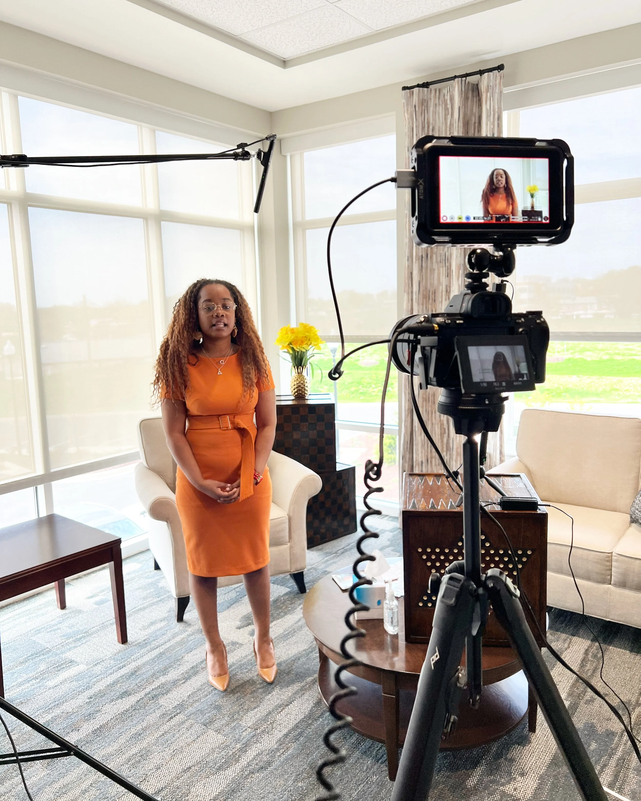 A woman in an orange dress and beige heels stands in a living room with large windows, decorated with a yellow flower in a gold vase on a side table, in front of a camera capturing her.