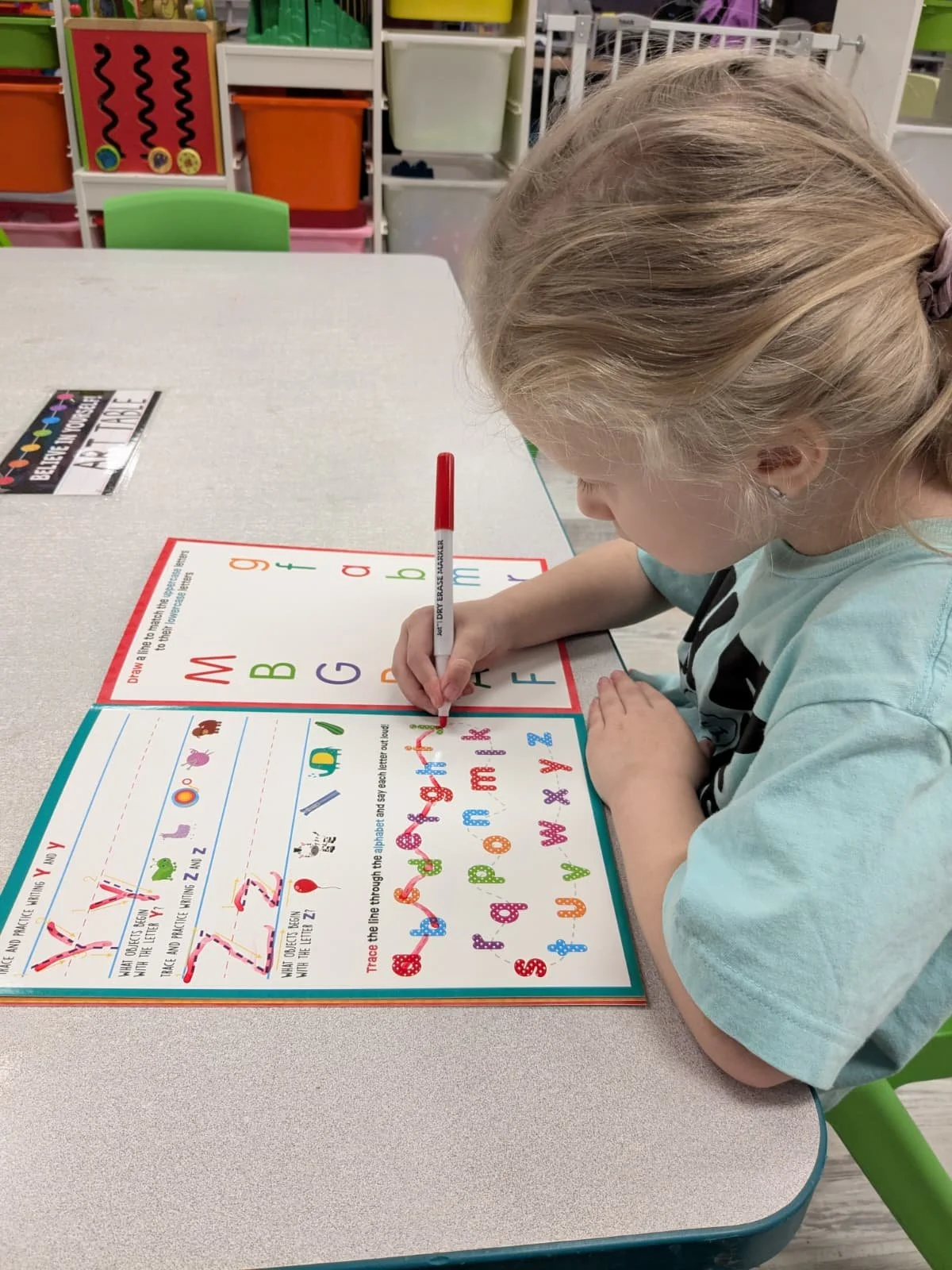 A young girl with blonde hair tied back, wearing a light blue t-shirt, is sitting at a table and tracing colorful alphabet letters in a workbook.