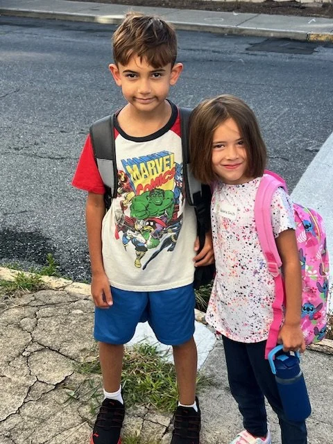 Two young children, a boy and a girl, standing on a sidewalk with backpacks, smiling at the camera.