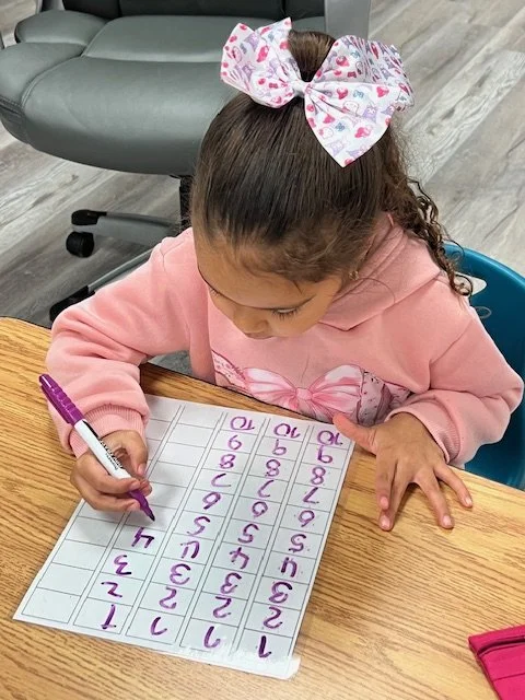 A young girl with curly hair in a high ponytail secured with a large pink and white bow is sitting at a wooden table, writing numbers on a whiteboard with a purple marker. She is wearing a pink sweatshirt and is focused on her activity.