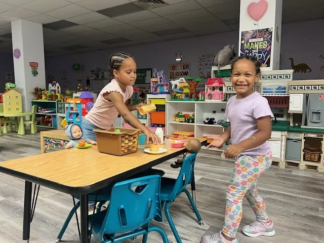 Two young girls playing with toys at a play kitchen set in a colorful indoor playroom.