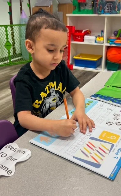 A young boy sitting at a table in a classroom, coloring in a workbook with an orange pencil.
