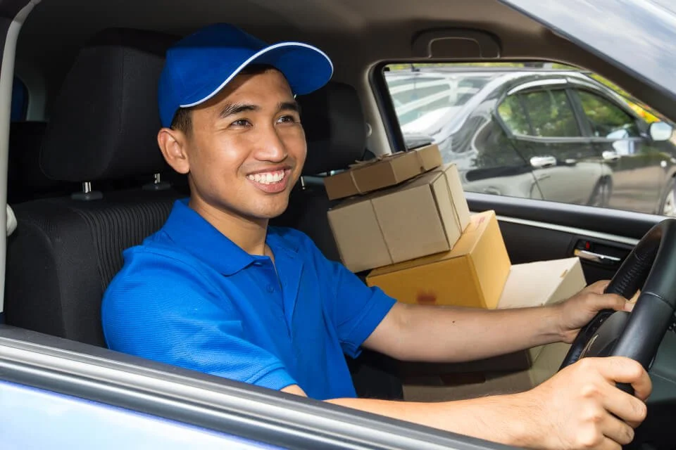 A smiling delivery person in a blue uniform and cap sitting in a car, holding the steering wheel, with boxes of packages in the back seat.