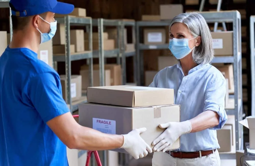 A woman and a man wearing masks and gloves exchanging boxes in a warehouse, with shelves of boxes in the background.