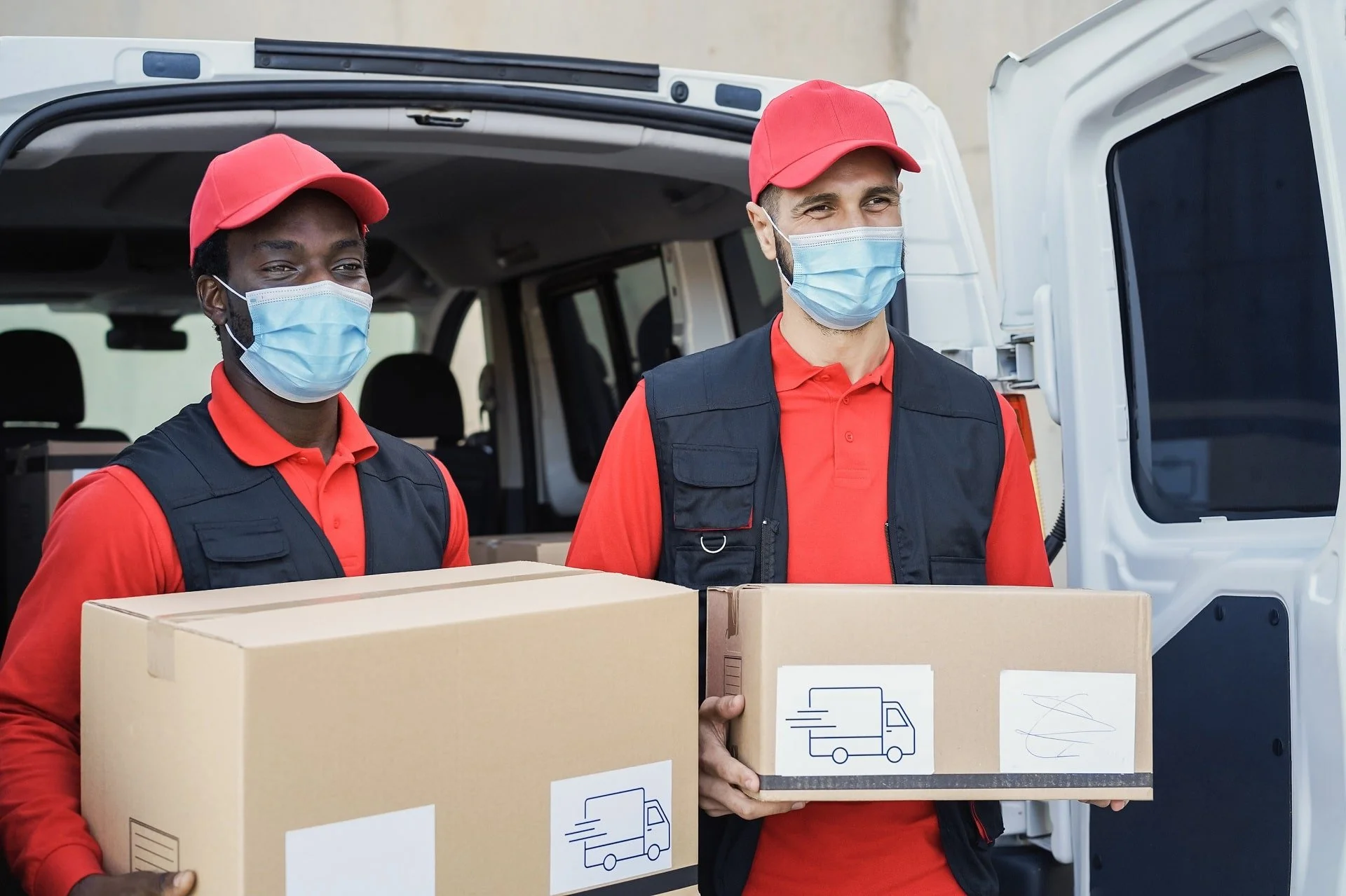 Two delivery workers wearing red caps and masks, holding cardboard boxes with delivery truck icons, standing near the open door of a delivery van.