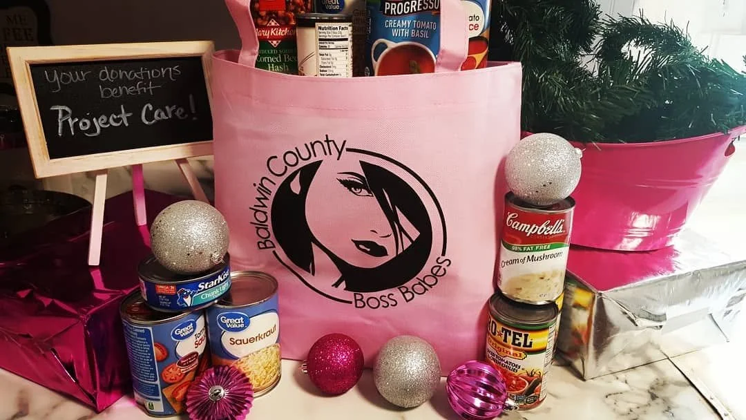Table display of canned soups, a pink tote bag with "Baldwin County Boss Babes" logo, pink and silver holiday ornaments, and a chalkboard sign that says "Your donation benefit Project Care!" in a festive setting.