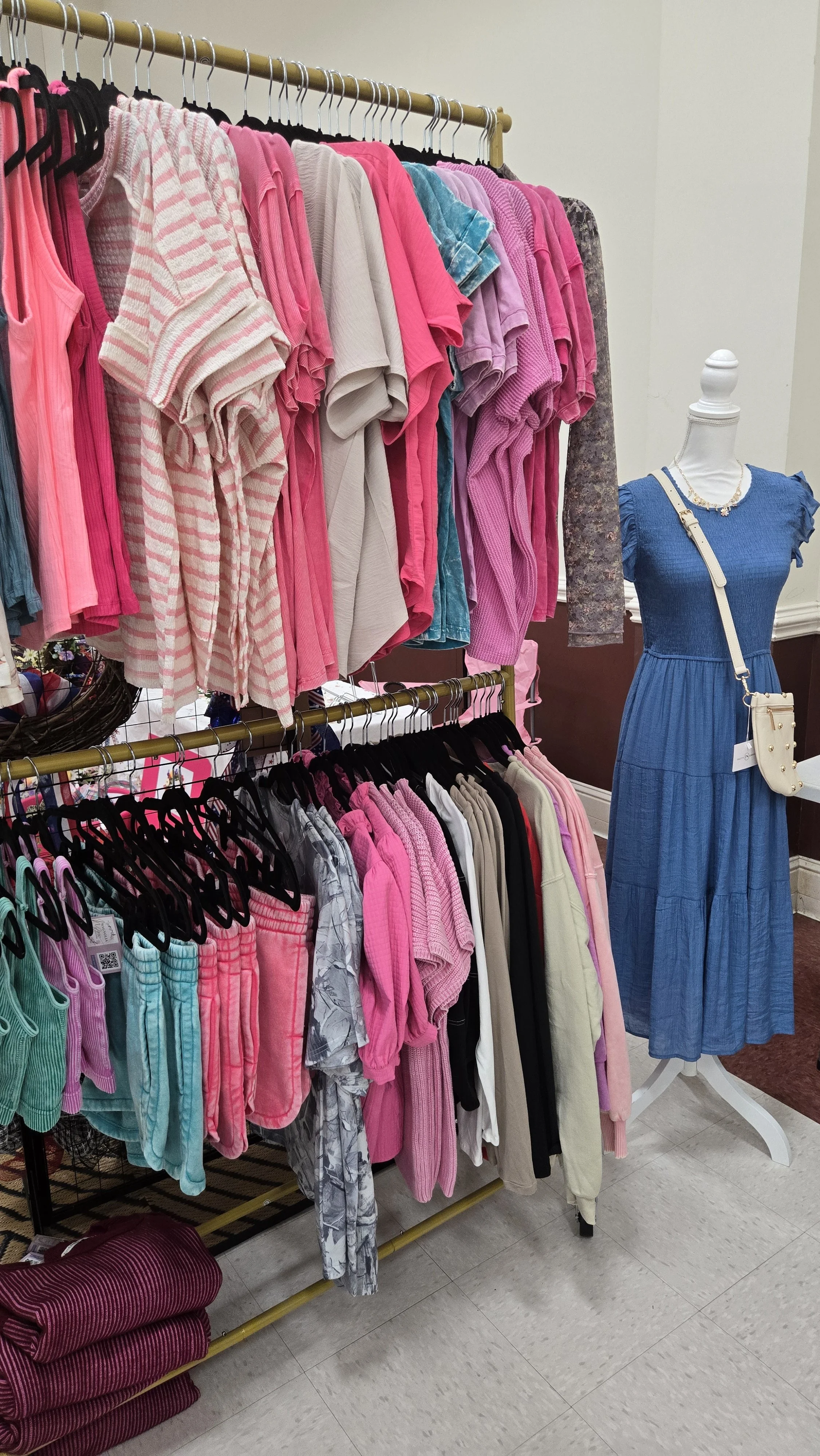 Clothing display with pink, white, turquoise, purple, and black garments on hangers and shelves, including a mannequin wearing a blue dress with a crossbody bag and jewelry.