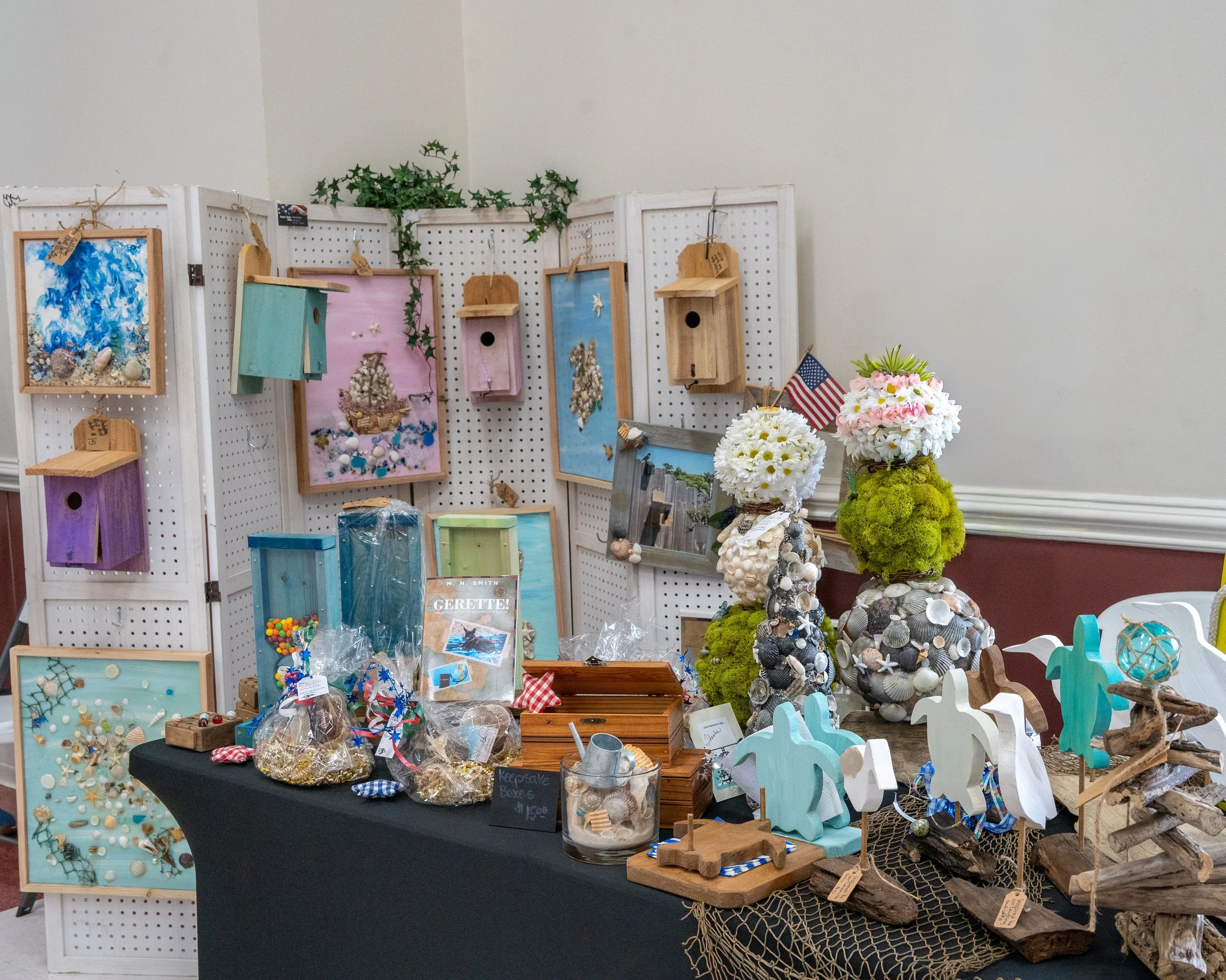 Display of handcrafted seashell and beach-themed decorations, photo frames, small birdhouses, and decorative objects at a craft fair or market.