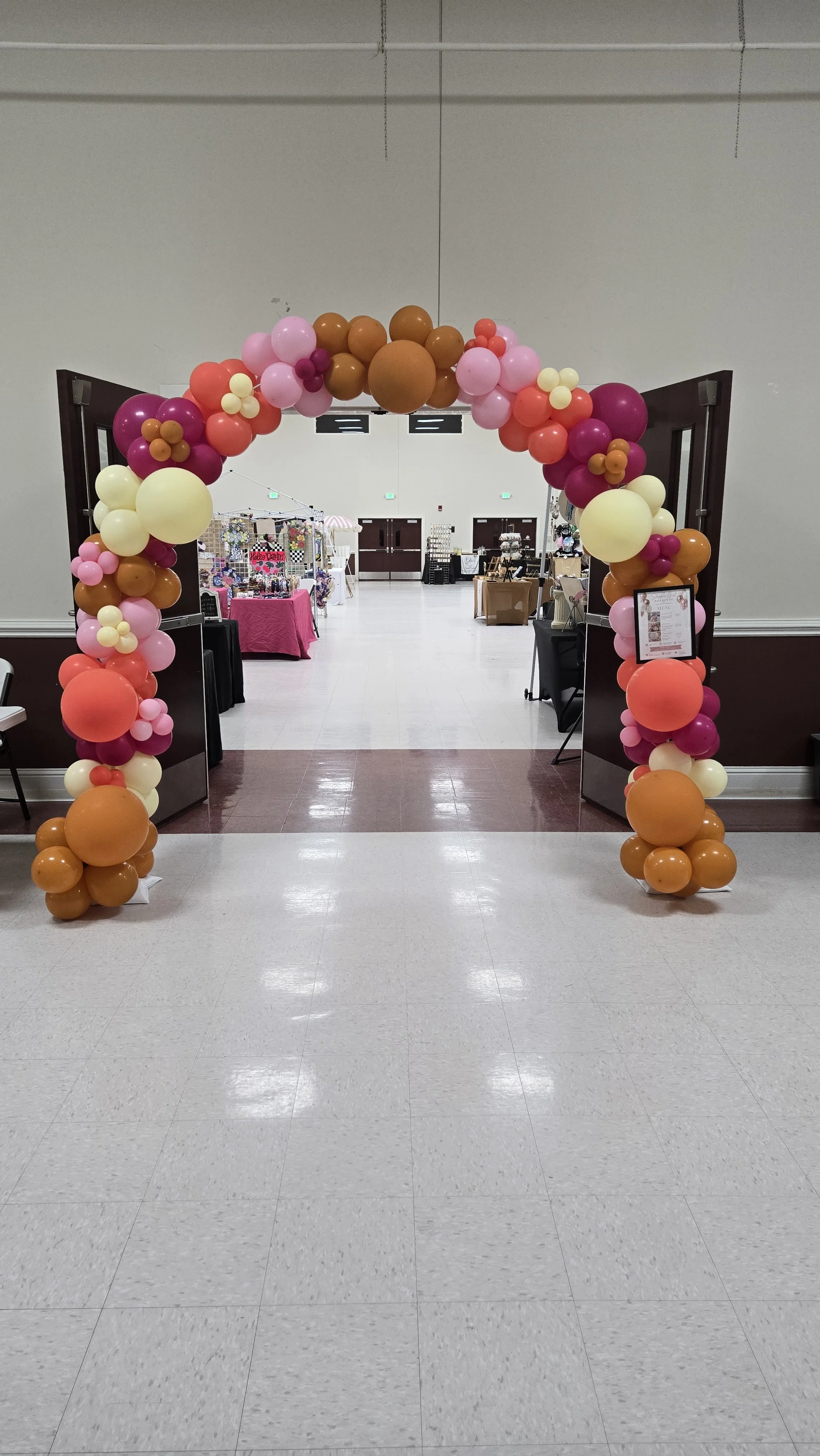 Balloon arch in pink, white, purple, orange, and cream colors at the entrance of an event space with vendor booths inside.