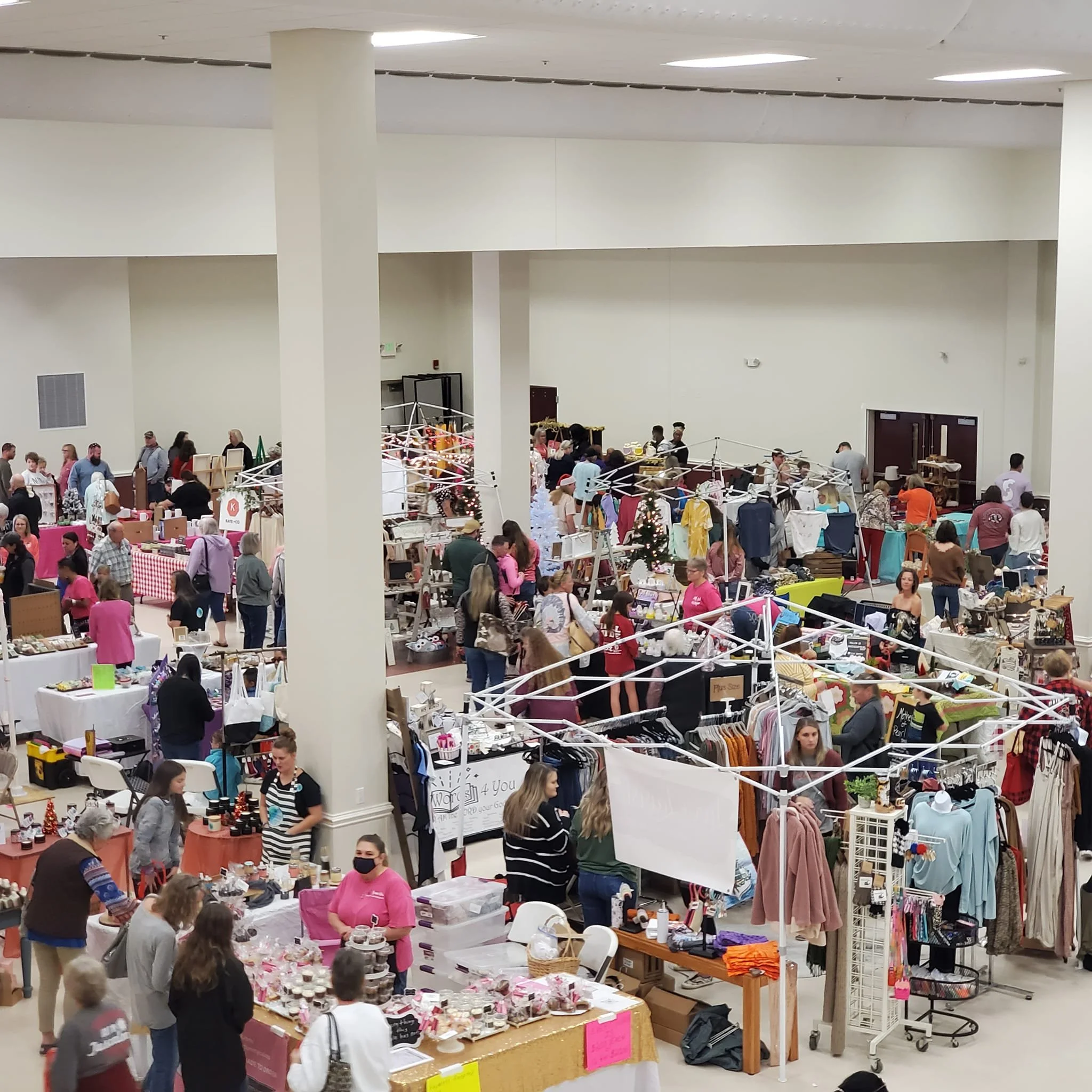 People shopping at a large indoor market or craft fair with various booths displaying items like clothing, accessories, and handmade crafts.