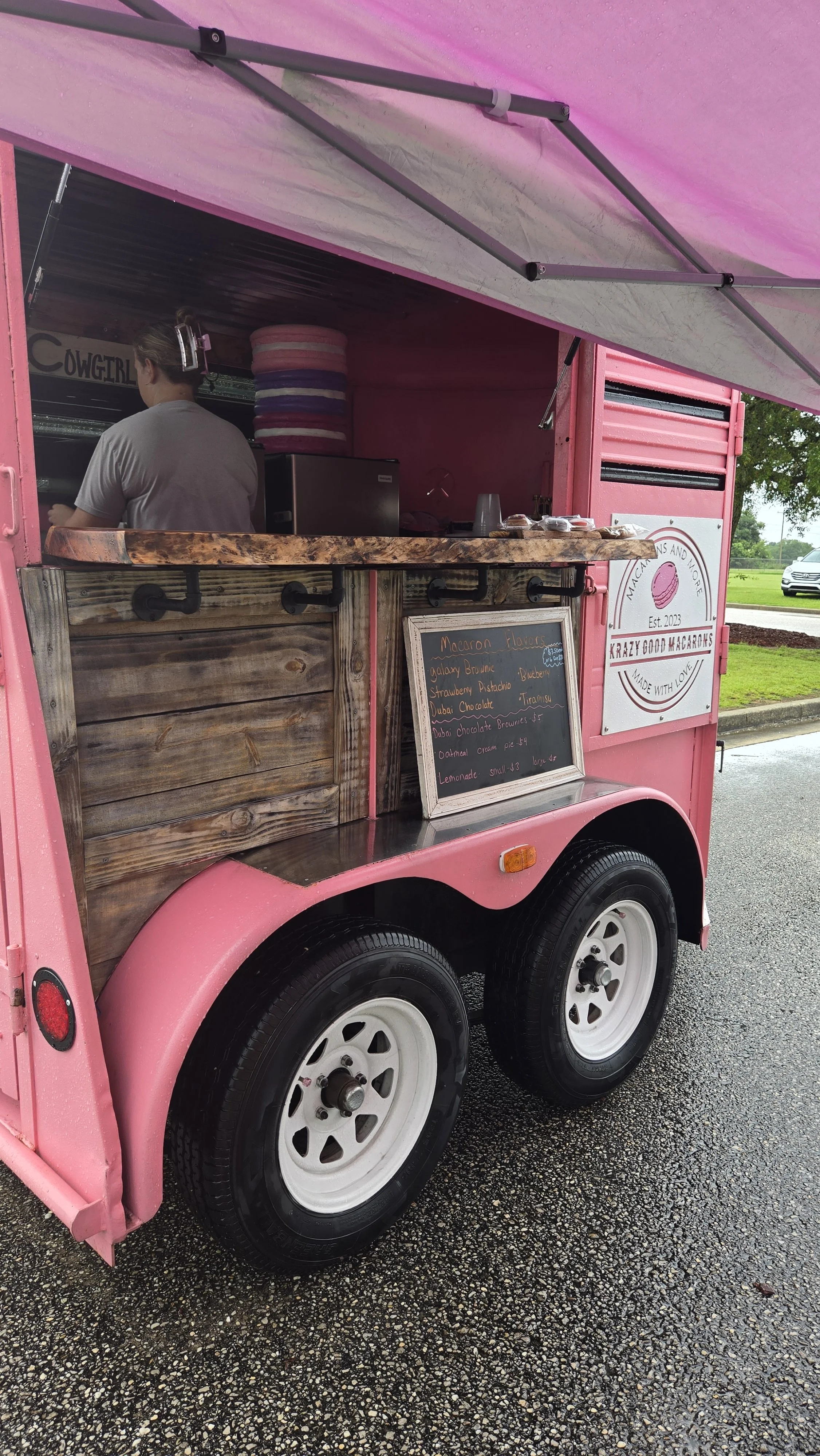 Pink mobile food stand selling macarons, with a blackboard menu listing flavors like galaxy brownie, strawberry pistachio, Dubai chocolate, and tiramisu, next to a grassy area and a tree.