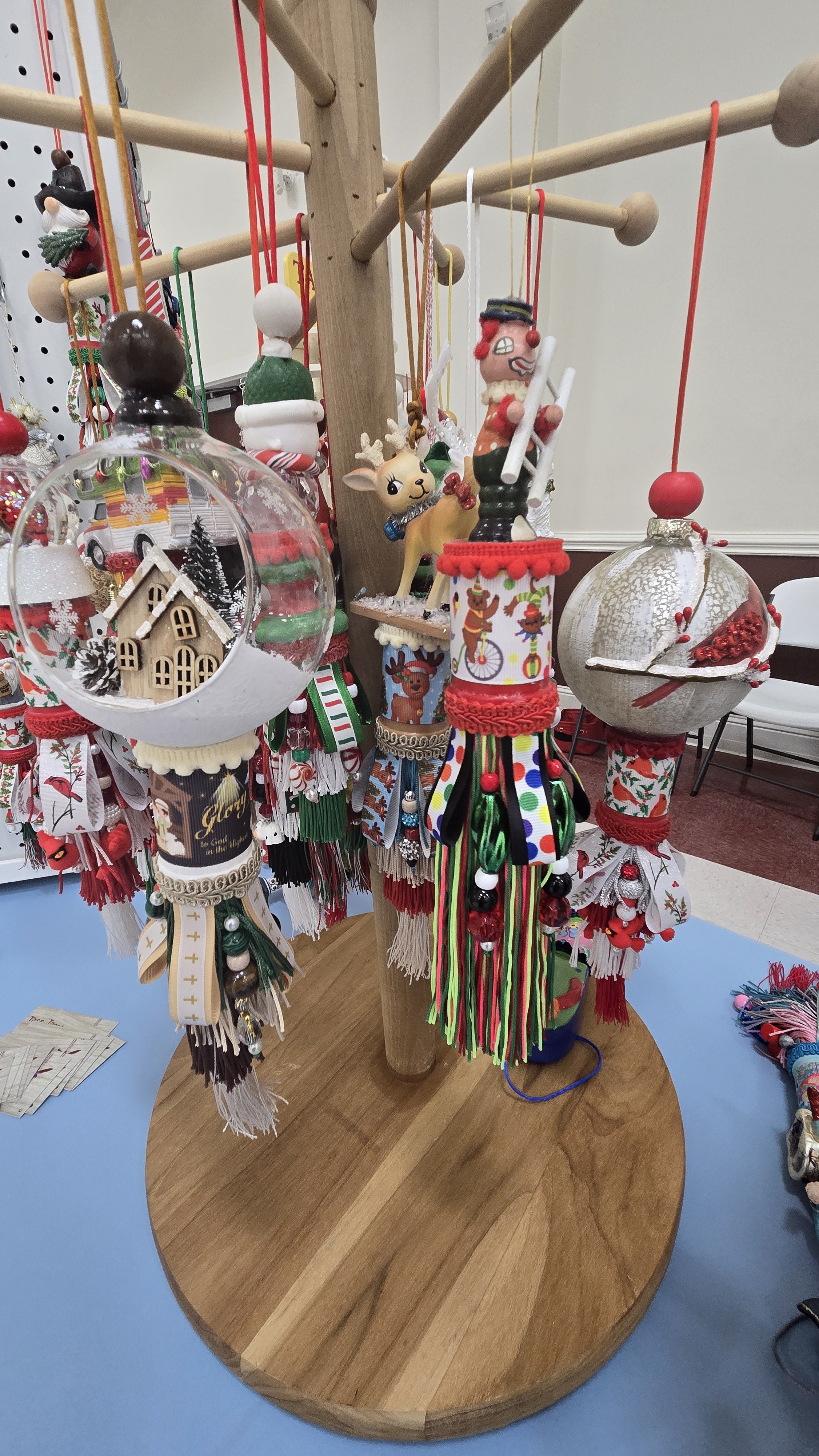 A wooden display stand holding Christmas ornaments, including decorated balls and figurines, with a blue table surface and chairs in the background.